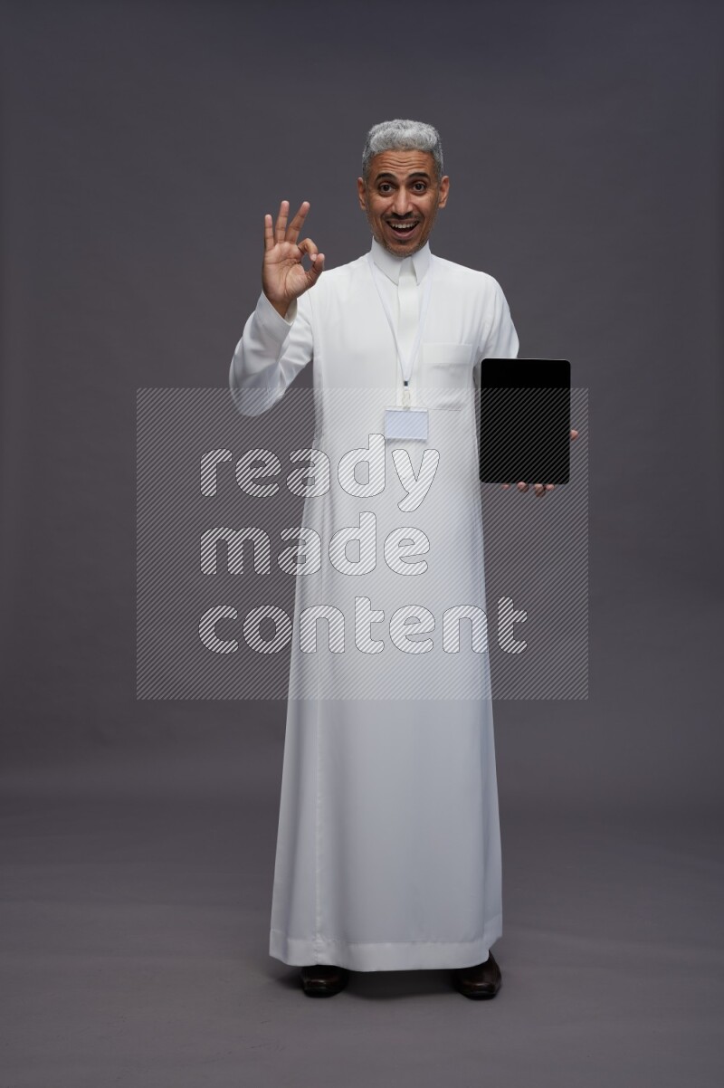 Saudi man wearing thob with neck strap employee badge standing showing tablet to camera on gray background