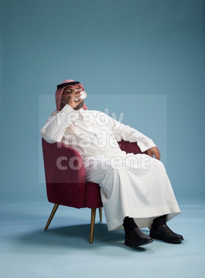 Saudi Man with shimag sitting on chair drinking coffee on blue background