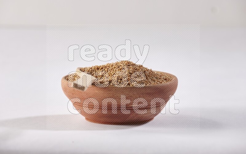 A wooden bowl full of mustard seeds with a wooden spoon on the seed on a white flooring