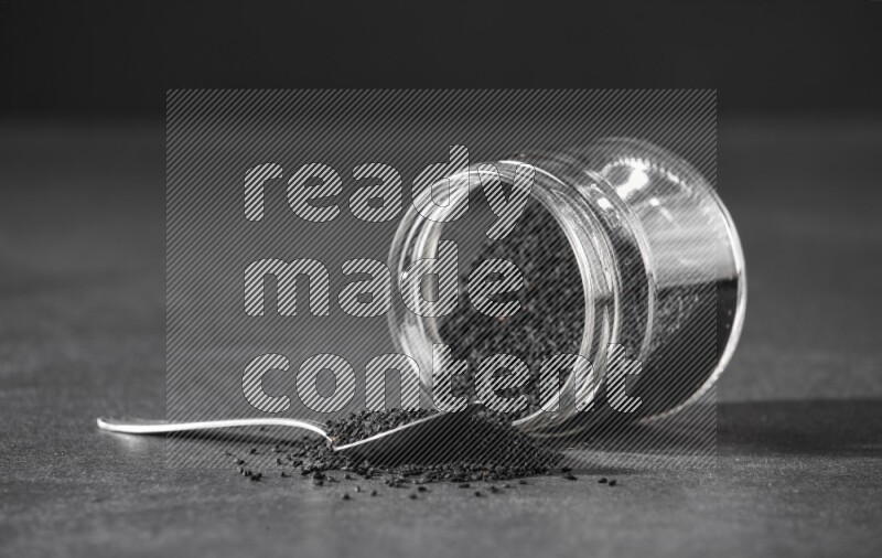 A flipped glass jar full of black seeds and seeds spread out with a metal spoon full of the seeds on a black flooring