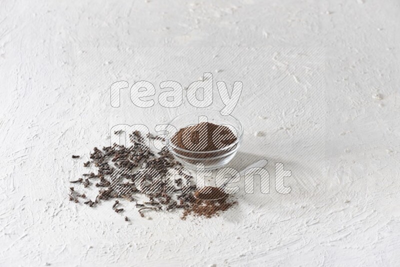 A glass bowl and a metal spoon full of cloves powder with cloves grains spread on a textured white flooring