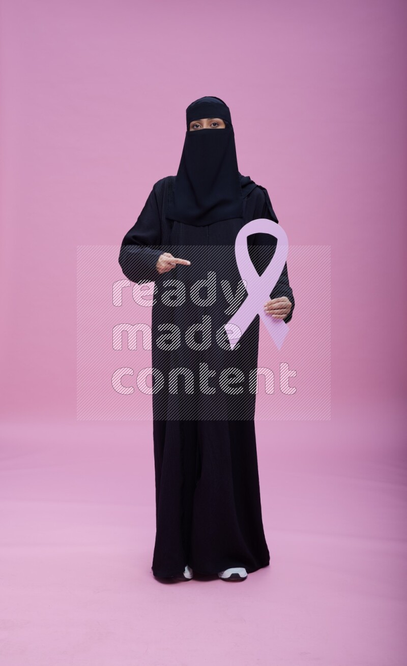 Saudi woman wearing abaya and niqab standing holding awareness ribbon on pink background
