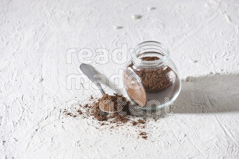 A glass spice jar and a metal spoon full of cloves powder on textured white flooring