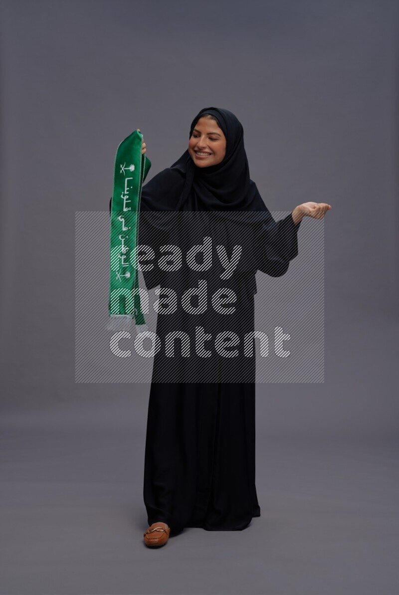 Saudi woman wearing Abaya standing holding Saudi flag on gray background