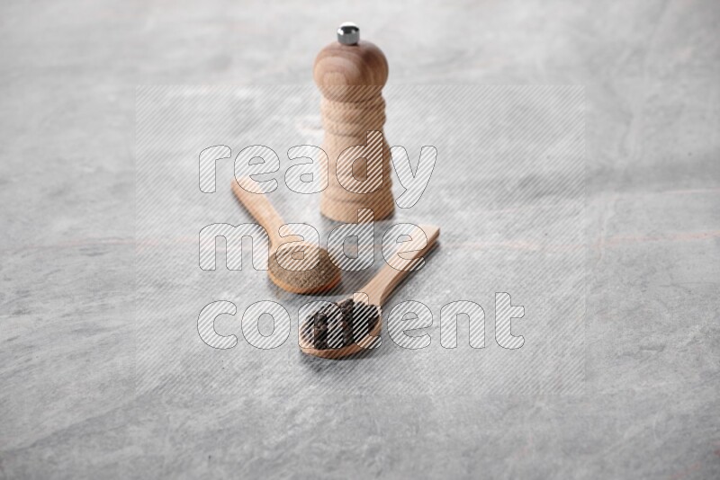 ملعقتان خشبيتان، ملعقة ممتلئة بمسحوق الفلفل الأسود والأخرى ممتلئة بحبات الفلفل الأسود ومطحنة خشبية على خلفية رخام رمادية