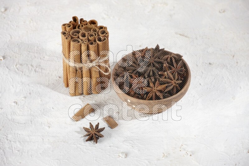 A stacked and bounded cinnamon sticks and a wooden bowl full of star anise on a white background
