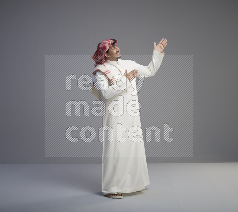 A Saudi man standing wearing thob and red shomag interacting with the camera on gray background