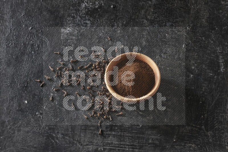 A wooden bowl full of cloves powder on a textured black flooring