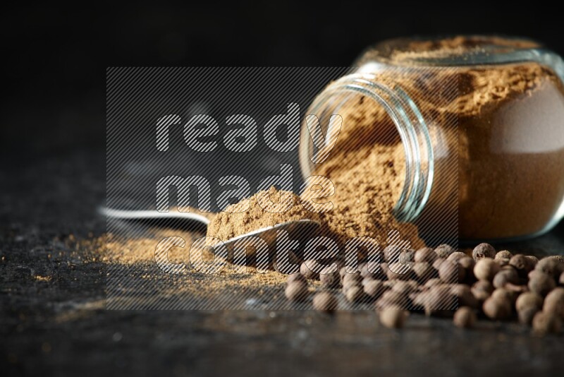 وعاء زجاجي للتوابل مقلوب وملعقة معدنية ممتلئان ببودرة بهار الفلفل الحلو والبودرة تناثرت من الوعاء مع بعض الحبوب على خلفية سوداء