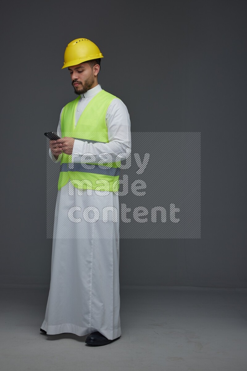 A Saudi man wearing Thobe with a yellow safety vest and white helmet standing and using his phone eye level on a gray background