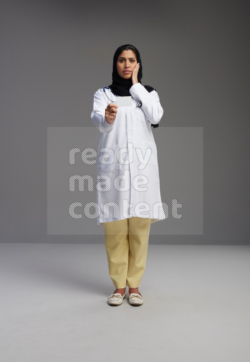 Saudi woman wearing lab coat with stethoscope standing holding ATM card on Gray background
