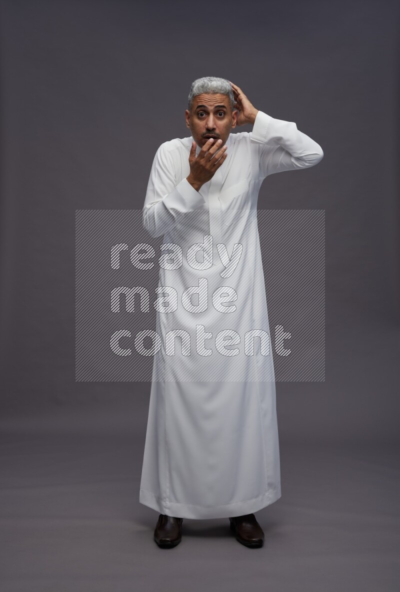 Saudi man wearing thob standing hands behind head on gray background