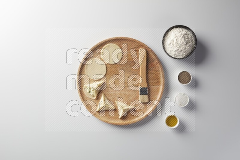 two closed sambosas and one open sambosa filled with cheese while flour, salt, black pepper and oil with oil brush aside in a wooden dish on a white background