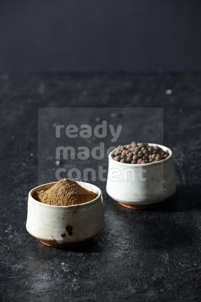 2 beige bowls, one full of allspice powder and the other full of whole balls on a textured black flooring