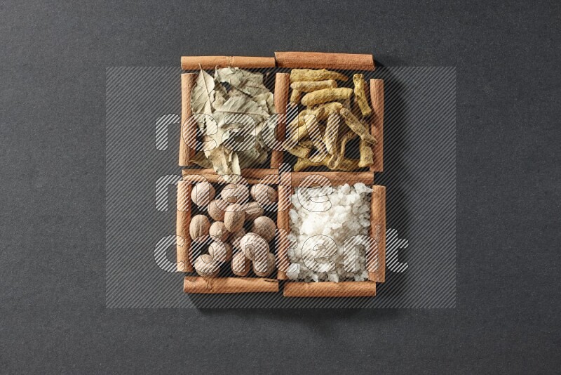 4 مربعات من عيدان القرفة ممتلئة بأوراق الغار والملح الأبيض وأصابع الكركم وجوزة الطيب على أرضية سوداء