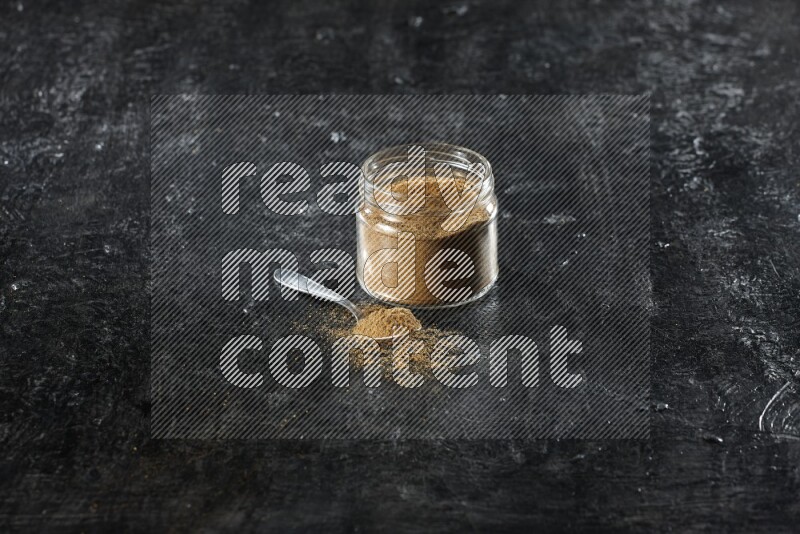 A glass jar and a metal spoon full of cumin powder on a textured black flooring