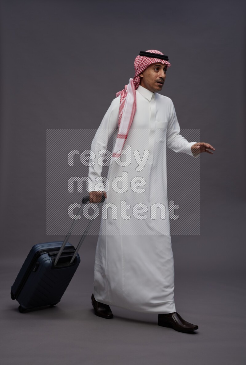 Saudi man wearing thob and shomag standing holding bag on gray background
