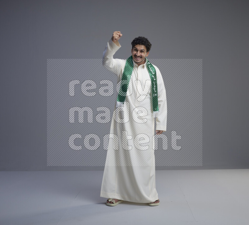 A Saudi man standing wearing thob and Saudi flag scarf on gray background