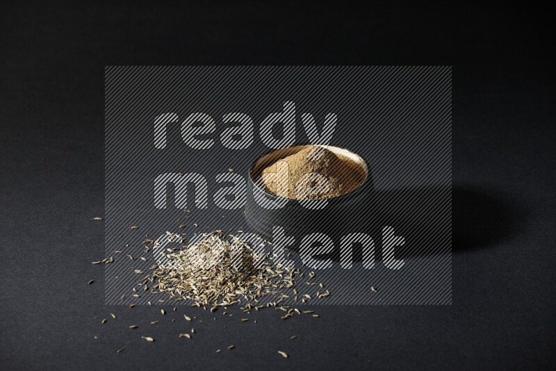 وعاء فخاري أسود ممتلئ ببودرة الكمون وحبوب الكمون متناثرة على خلفية سوداء