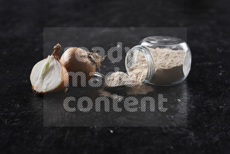 A glass jar full of onion powder flipped with some spilling powder on black background