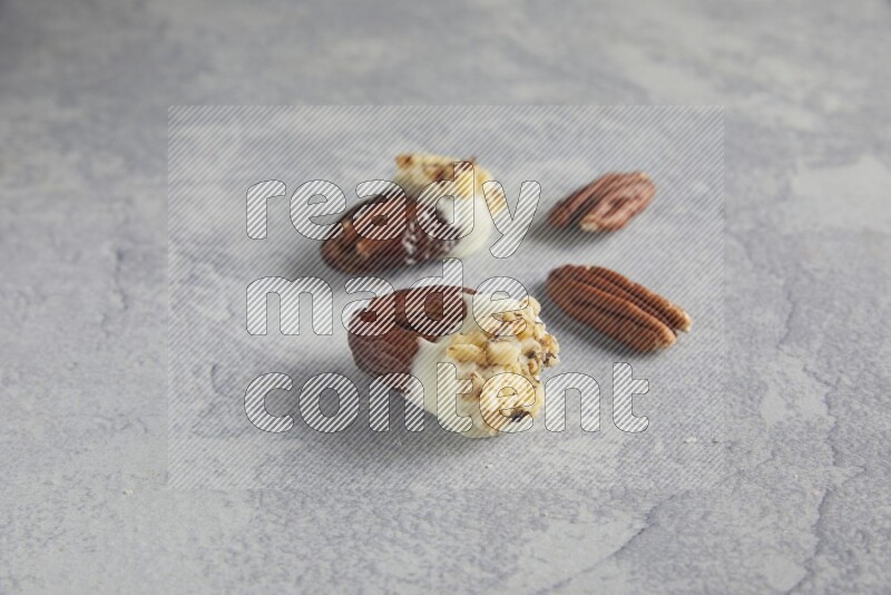 Two pecan stuffed dates covered with white chocolate and chopped walnuts with unroasted pecans on alight grey background