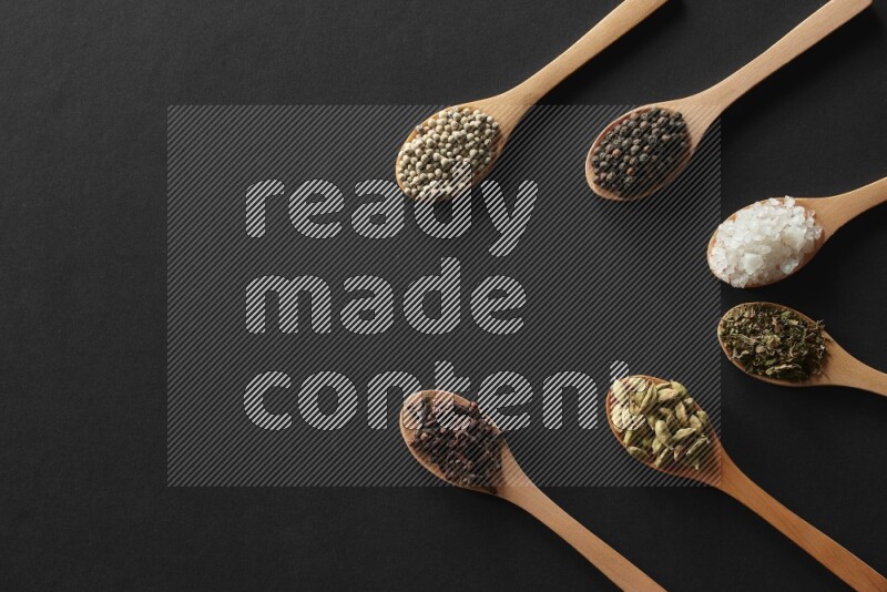 الفلفل الأبيض والقرنفل والهيل والملح والفلفل الأسود وريحان مجفف في معالق خشبية علي خلفية سوداء