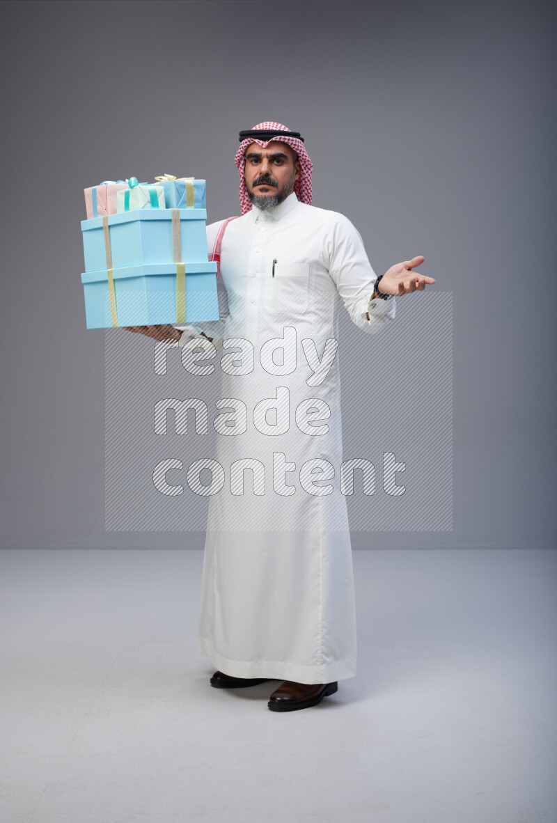 Saudi man Wearing Thob and red Shomag standing holding gift box on Gray background