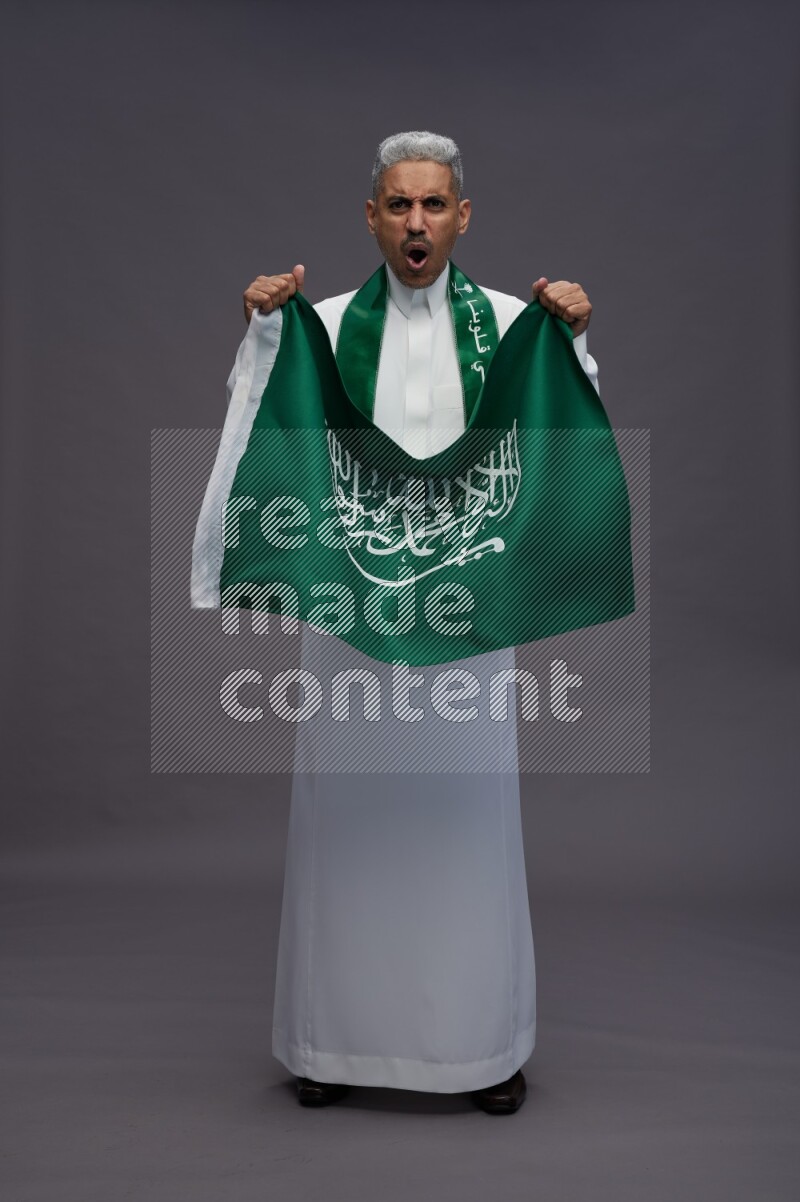 Saudi man wearing thob standing holding Saudi flag on gray background