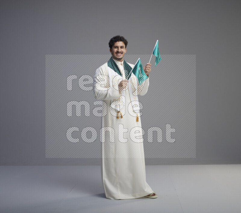 A Saudi man standing wearing thob and saudi flag scarf and holding small saudi flag on gray background