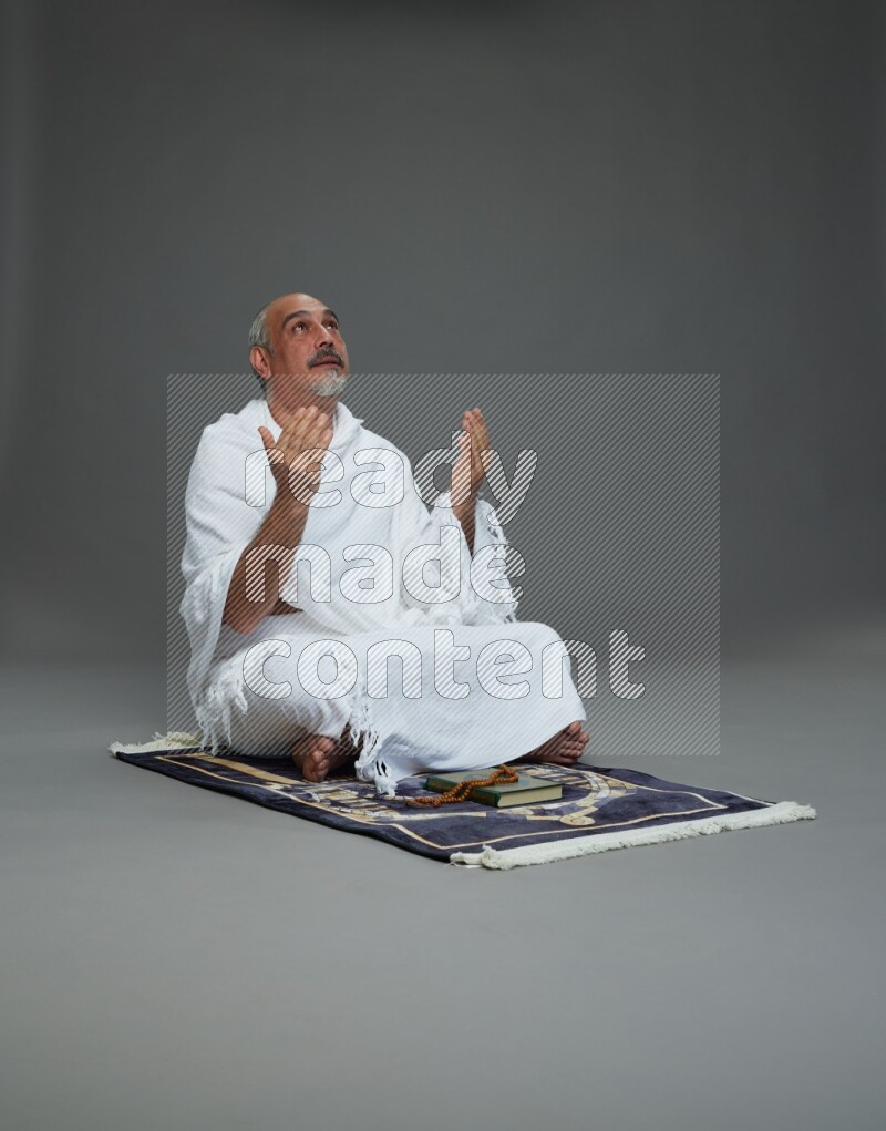 A man wearing Ehram sitting on prayer mat dua'a on gray background