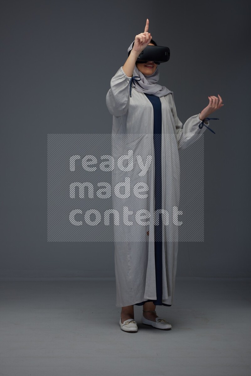 A Saudi woman wearing a light gray Abaya and head scarf standing and wearing VR eye level on a grey background