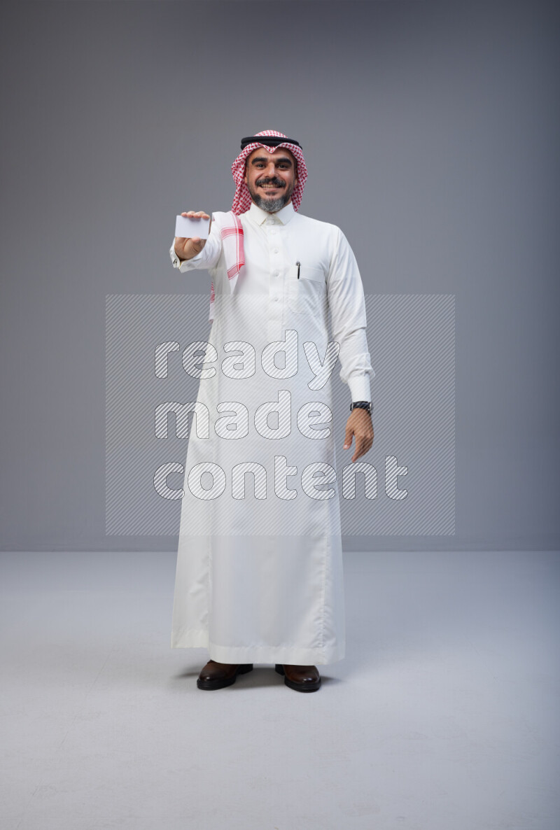 Saudi man Wearing Thob and red Shomag standing holding ATM card on Gray background