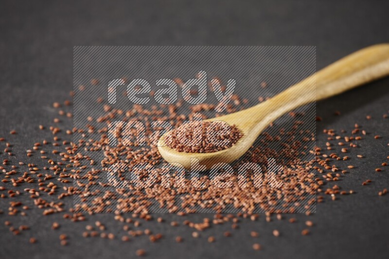 A wooden spoon full of garden cress seeds and seeds spread beside it on a black flooring
