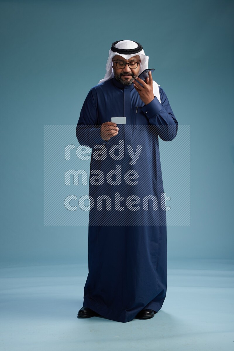 Saudi Man with shimag Standing holding ATM card while talking on phone on blue background