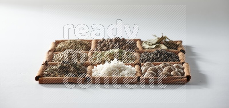 9 squares of cinnamon sticks full of cardamom in the middle surrounded by nutmeg, salt, dried basil, white pepper, cumin, allspice, bay laurel leaves and black pepper on white flooring