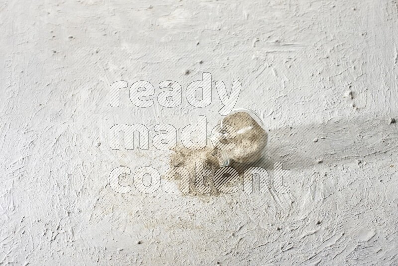 A flipped herbal glass jar full of white pepper powder with spilled powder on textured white flooring