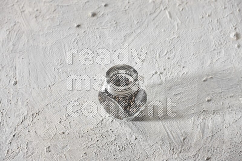 A glass spice jar full of black pepper on textured white flooring