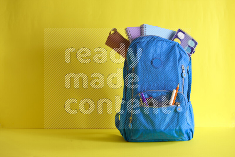 A school bag with assorted school supplies in and beside it on yellow background