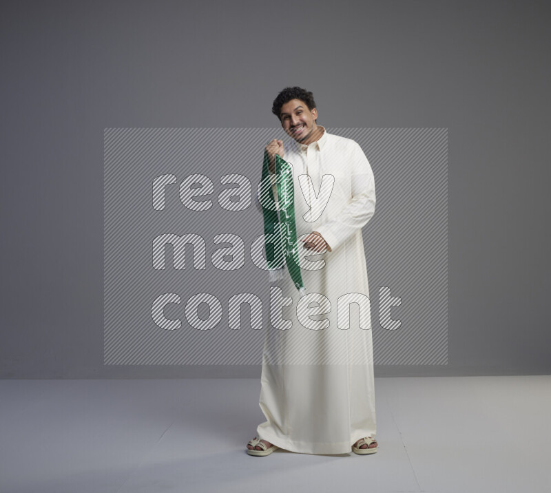 رجل سعودي يرتدي ثوب ابيض ويحمل شال اخضر على خلفية رمادي