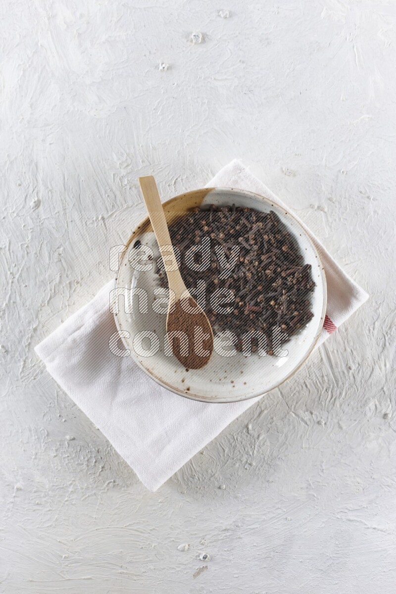 A Pottery plate full of whole cloves and a wooden spoon full of cloves powder in it on a textured white background