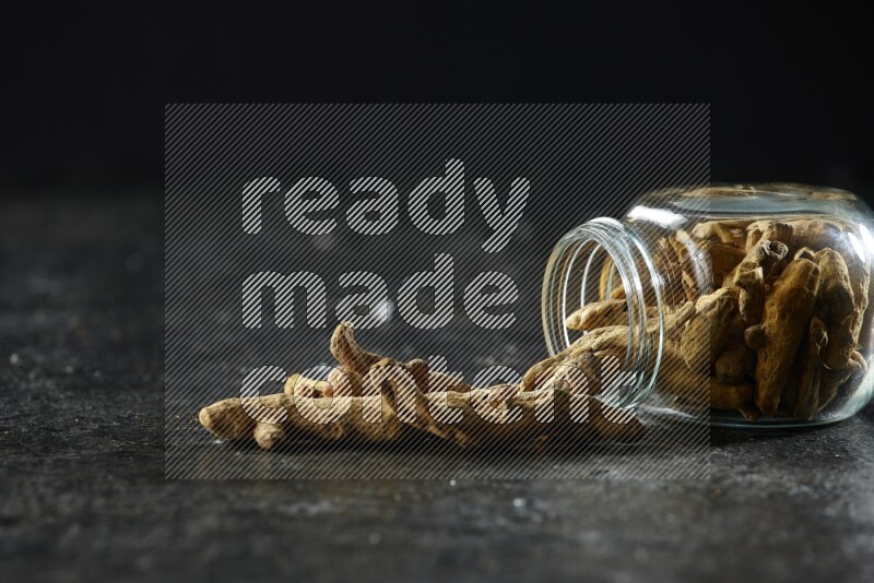 وعاء زجاجي للتوابل مقلوب وممتلئ بأصابع الكركم المجففة وبعض أصابع الكركم خارج الوعاء على خلفية سوداء