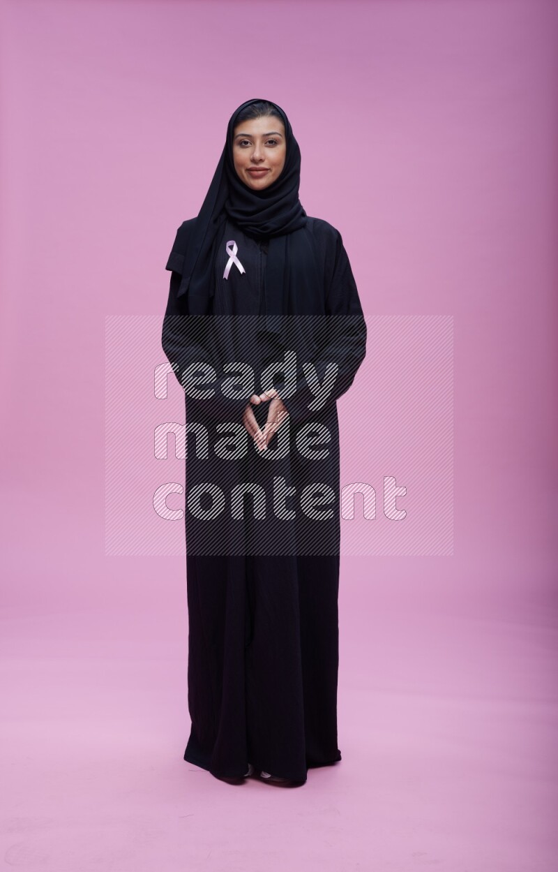 Saudi woman wearing pink ribbon on Abaya standing interacting with the camera on pink background