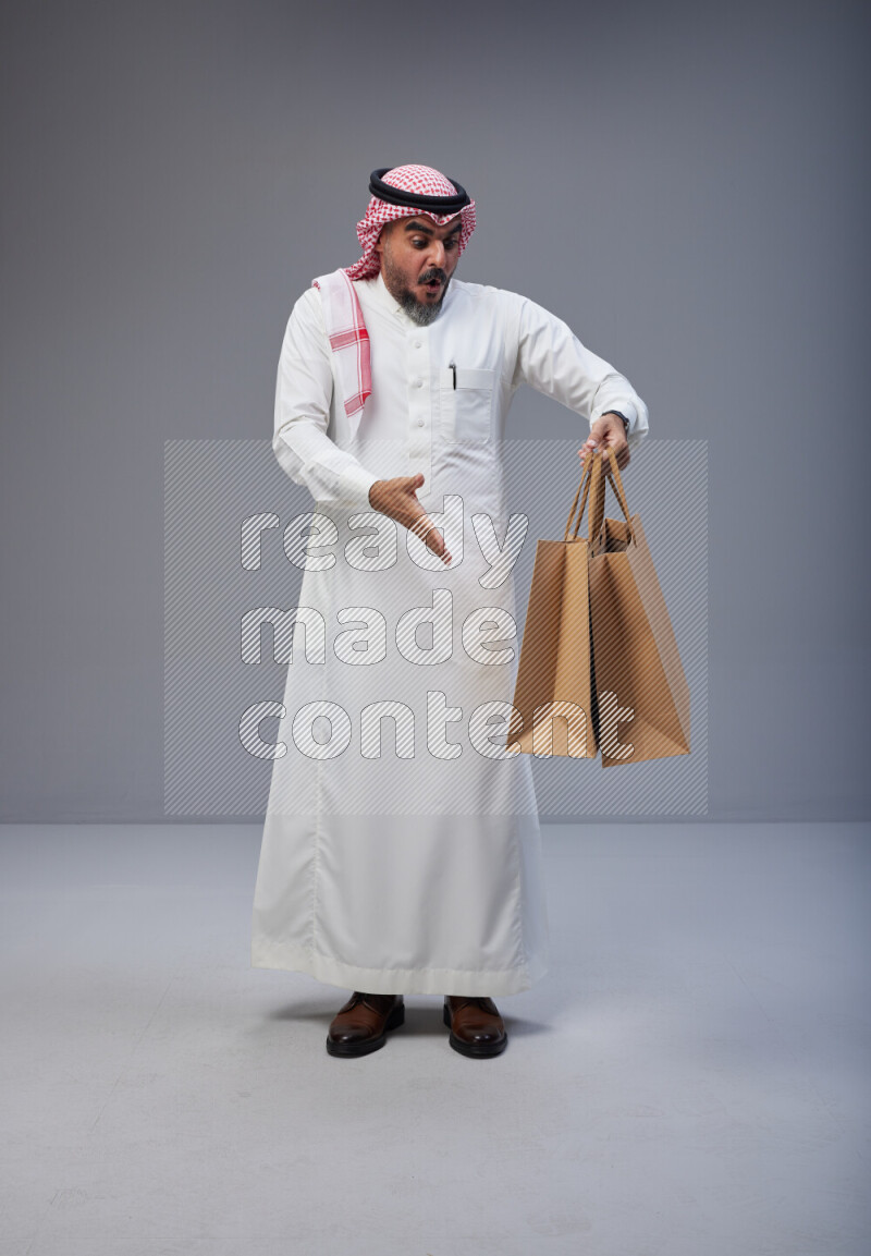 Saudi man Wearing Thob and red Shomag standing holding shopping bag on Gray background