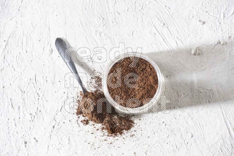A glass jar full of cloves powder with a metal spoon on a textured white flooring