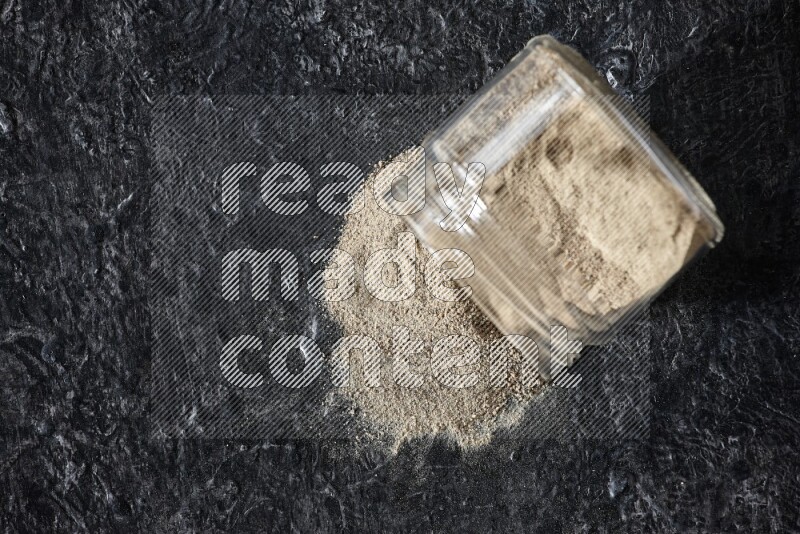 A flipped glass jar full of white pepper powder with spilled powder on textured black flooring
