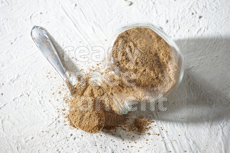 A flipped glass spice jar and a metal spoon full of cumin powder and powder spilled out on textured white flooring