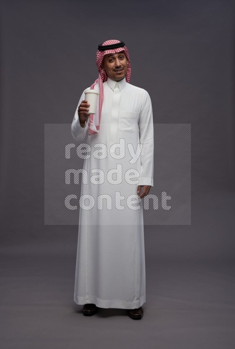 Saudi man wearing thob and shomag standing holding paper cup on gray background