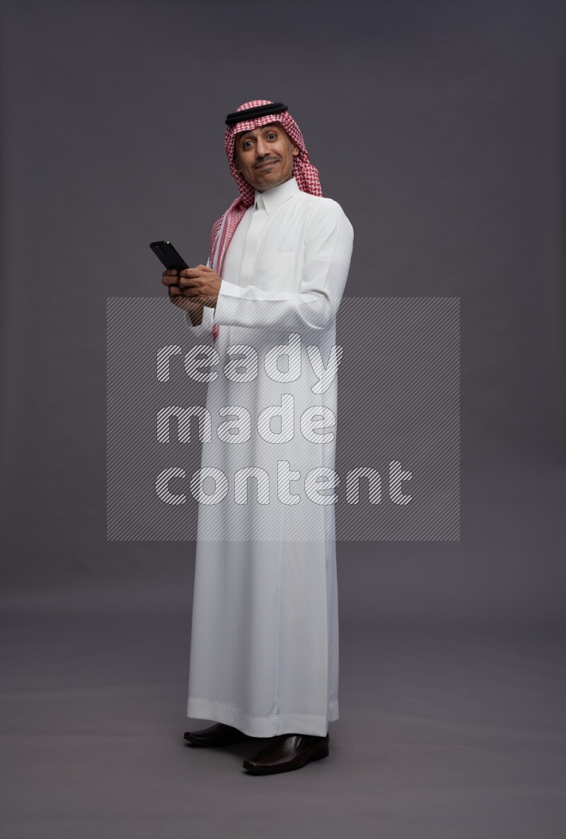 Saudi man wearing thob and shomag standing texting on phone on gray background