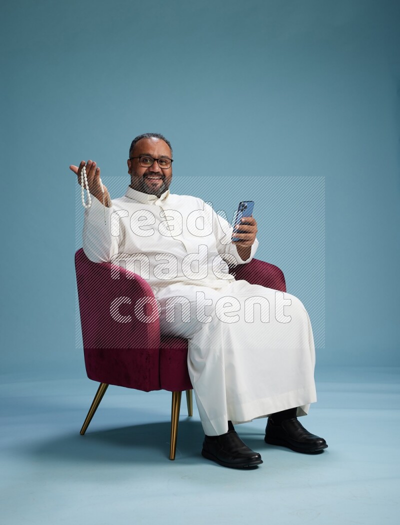 Saudi Man without shimag sitting on chair texting on phone on blue background