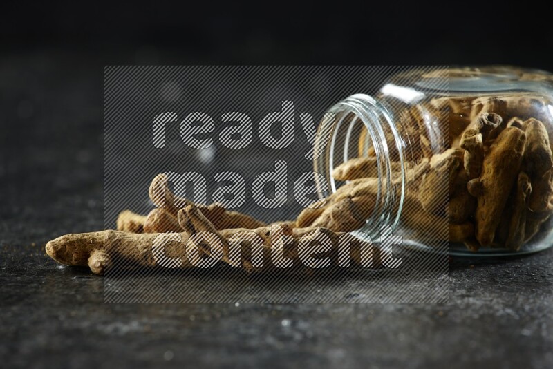 A flipped glass spice jar full of dried turmeric whole fingers and fingers fell out of it on textured black flooring